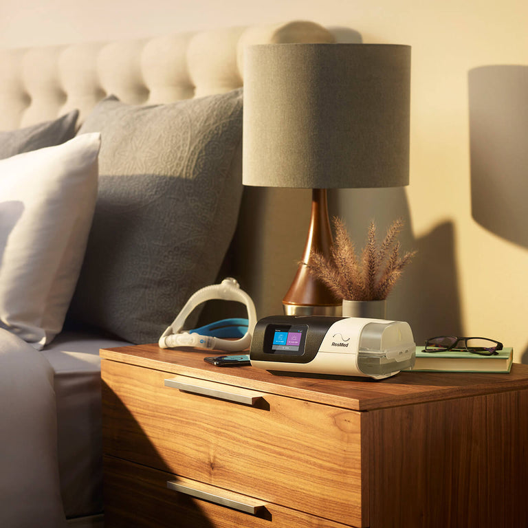 AirSense 11 CPAP Machine Autoset on top of a side table, next to the CPAP Machine is a ResMed CPAP Mask. It is next to a bed.